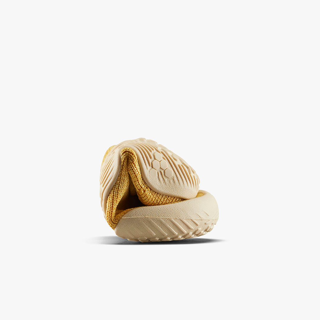 Fleksibel Vivobarefoot Pluma Knit Toddlers sko i Honey Gold, rullet for at vise dens bøjelighed med en hexagonal mønstret sål. Designet til barfodsfølelse og bevægelsesfrihed.