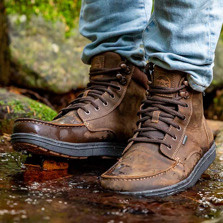 Lems Boulder Boot Grip Waterproof Dakota variant på en model, brun med snørebånd, velegnet til udendørsbrug og står i vandløb, hvilket viser deres vandtætte funktion.