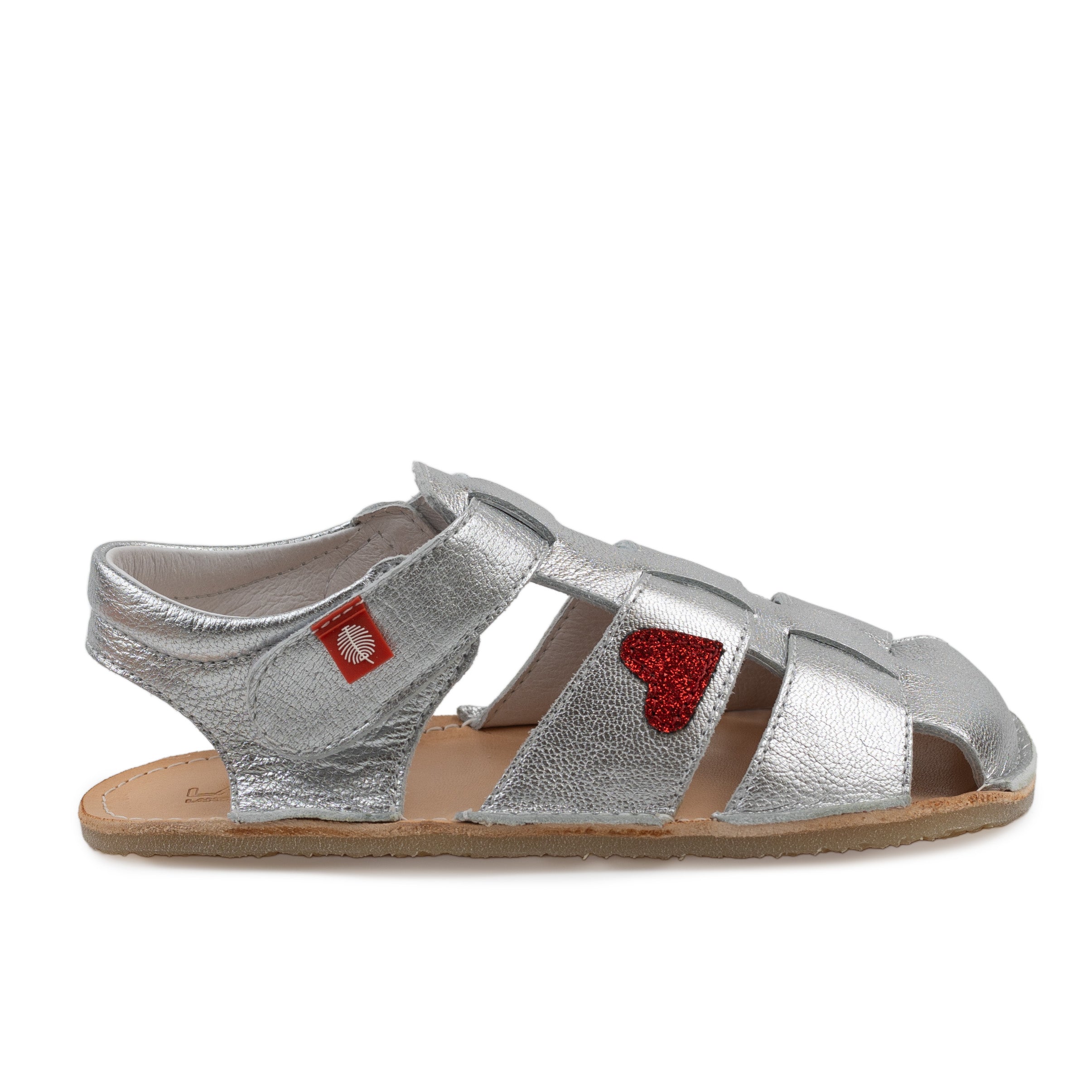 Tikki Lasta Leather Cuore bæresandal til børn i sølv med rød glitterhjerte. Unisexdesign med åben tå og velcrolukning, behagelig naturfarvet sål og mærketag.