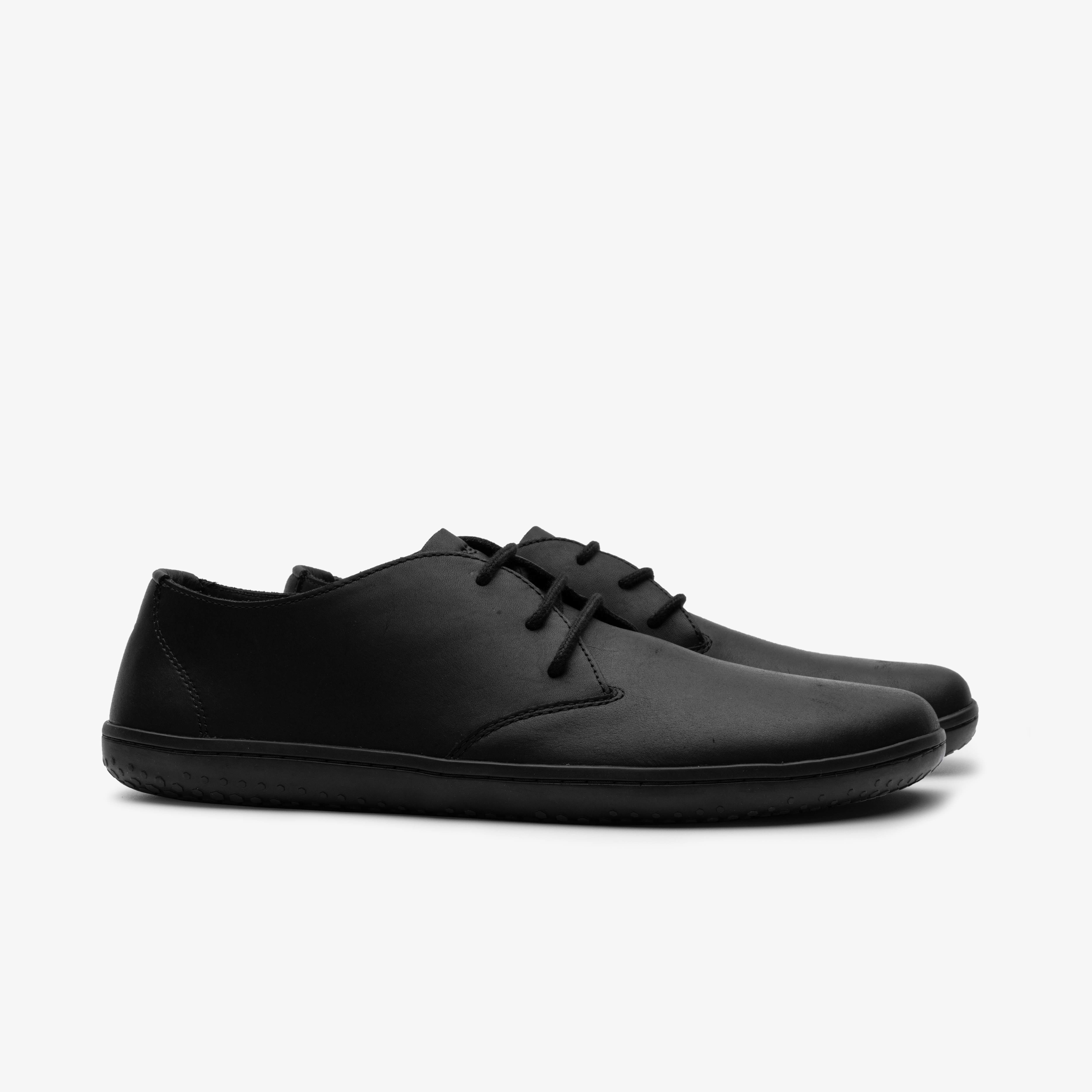 Par Vivobarefoot Ra IV i varianten Obsidian, minimalistisk designet barfodssko i sort vildtlæder med økologiske bomuldssnørebånd og en fleksibel ydersål, ideel til at fremme naturlig fodbevægelse.