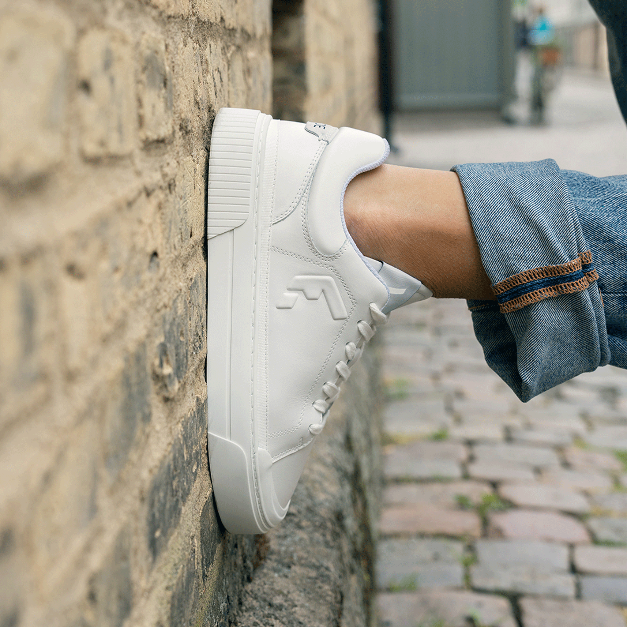 Unisex Wyde Arcus sneaker in all white, shown on foot against a brick wall, emphasizing its smooth leather finish and ribbed sole.