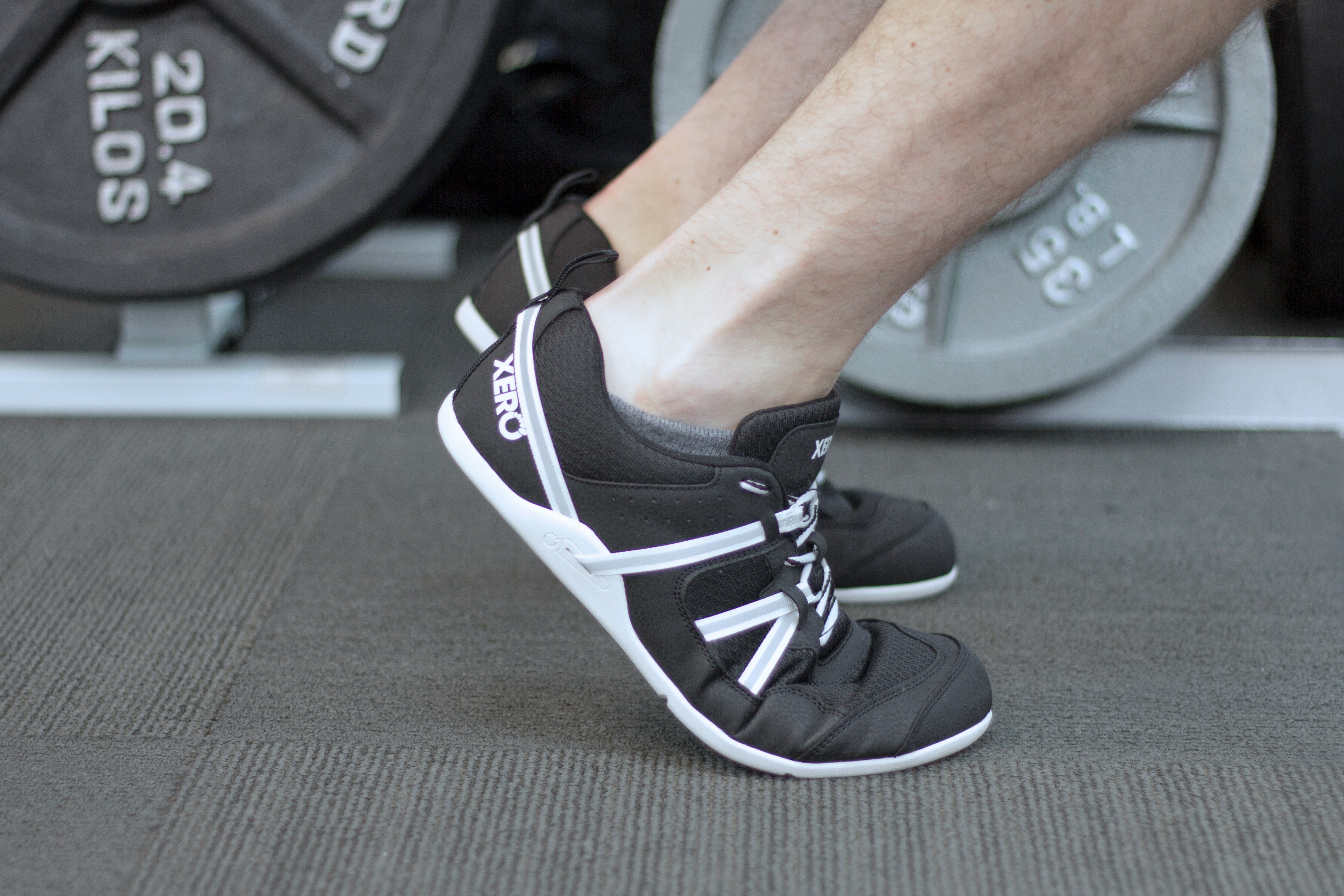 Mand iført Xero Shoes Prio Men barefoot sko i Black/White variant i et fitnesscenter. Skoene tilbyder naturlig pasform med mesh overdel og tynd, fleksibel sål til jordfornemmelse.